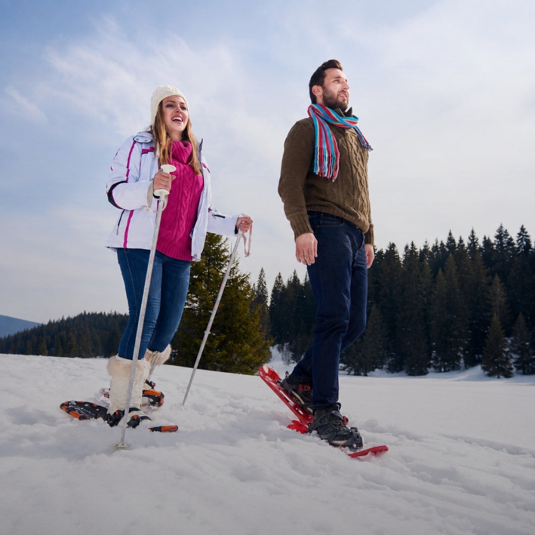 A man and a woman snowshoeing in a snowy outdoor setting with pine trees in the background. The woman wears a white and pink jacket, blue jeans, and white snow boots, while the man wears a brown sweater, dark jeans, and a colorful striped scarf.