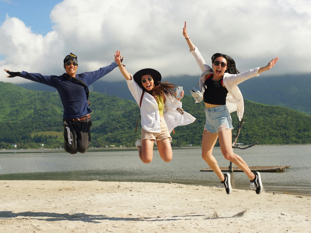 Three friends jumping excitedly on a sandy beach with water and green hills in the background. They are wearing casual summer outfits, including a navy long-sleeve top, white shirt and shorts, and a black top with a white cardigan.
