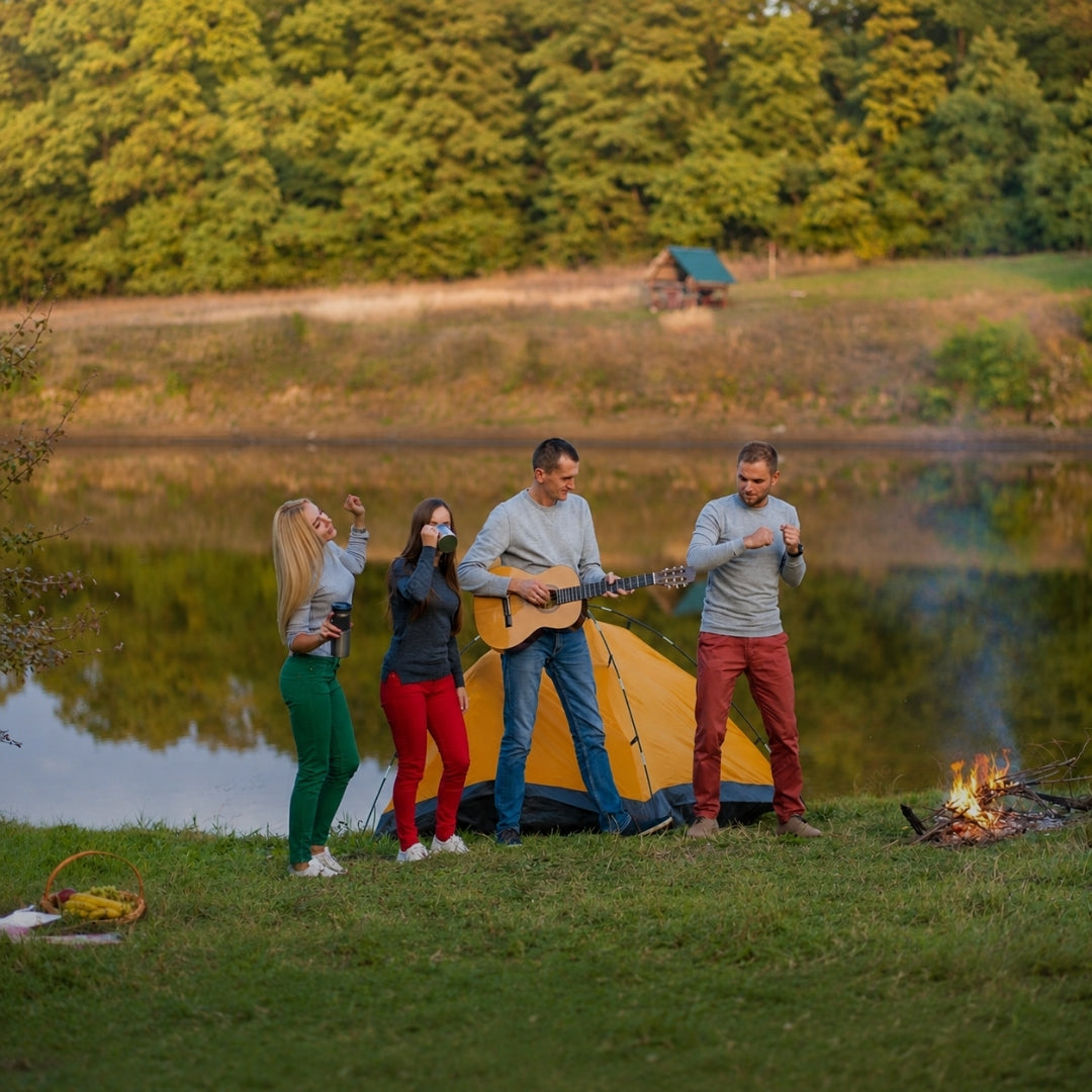 Four people camping by a lake at dusk, with a yellow tent, a small campfire, and a forest in the background. One person is playing an acoustic guitar, and the group appears to be singing or dancing.