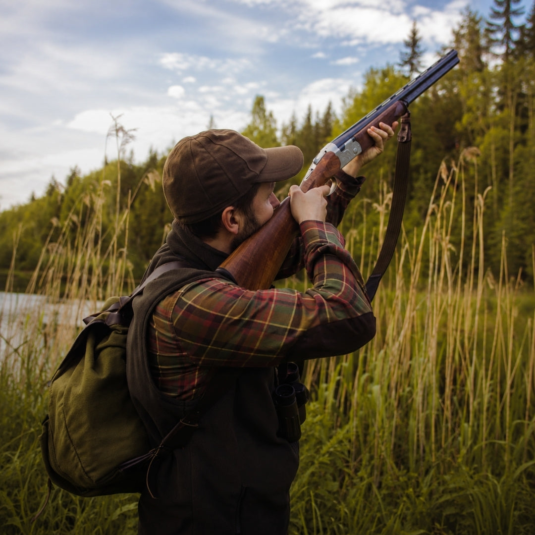 A man in outdoor hunting attire, including a brown cap, plaid shirt, and green backpack, aiming a double-barrel shotgun in a grassy field with trees and water in the background.